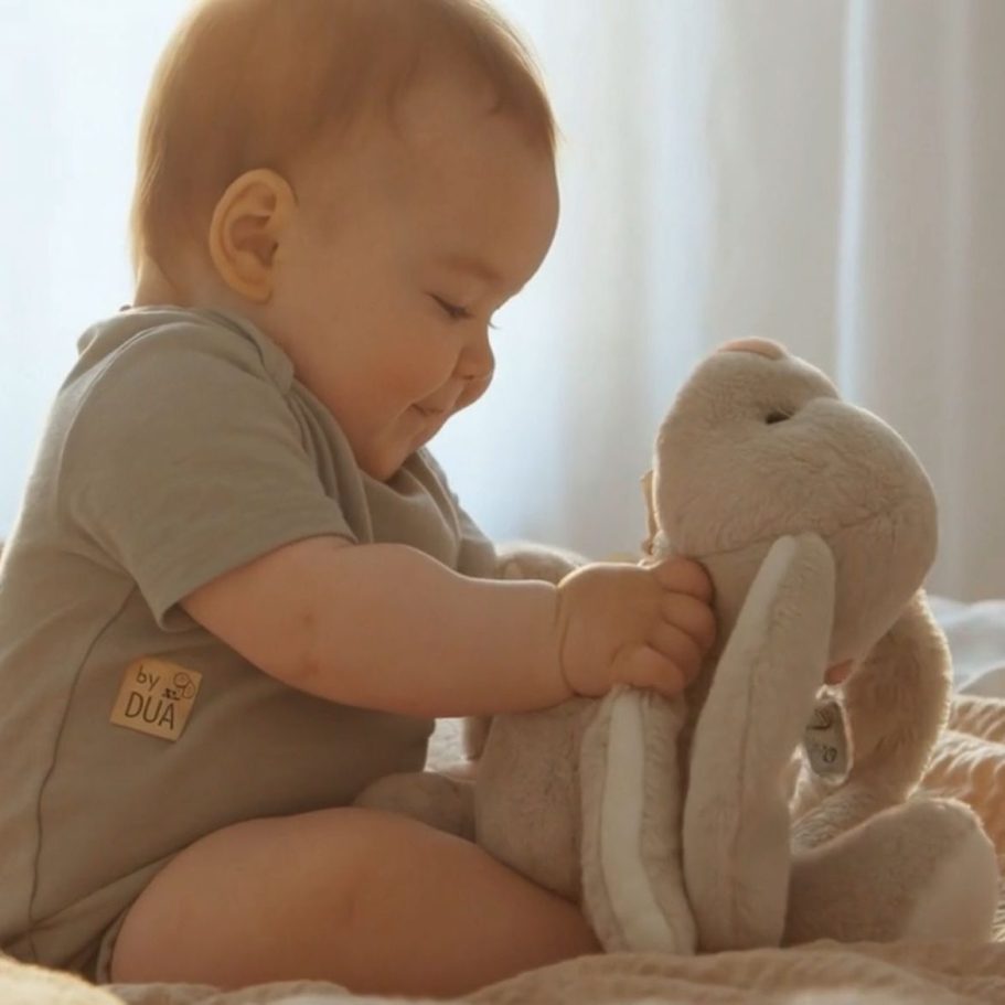 Lächelndes Baby, das mit einem plüschigen Hasen spielt. by DUA Babyartikel 